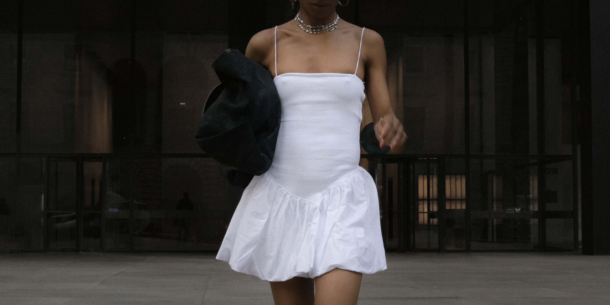 A woman in a white tennis dress, black sneakers, an oversized black tote bag, baseball cap, and silver jewelry