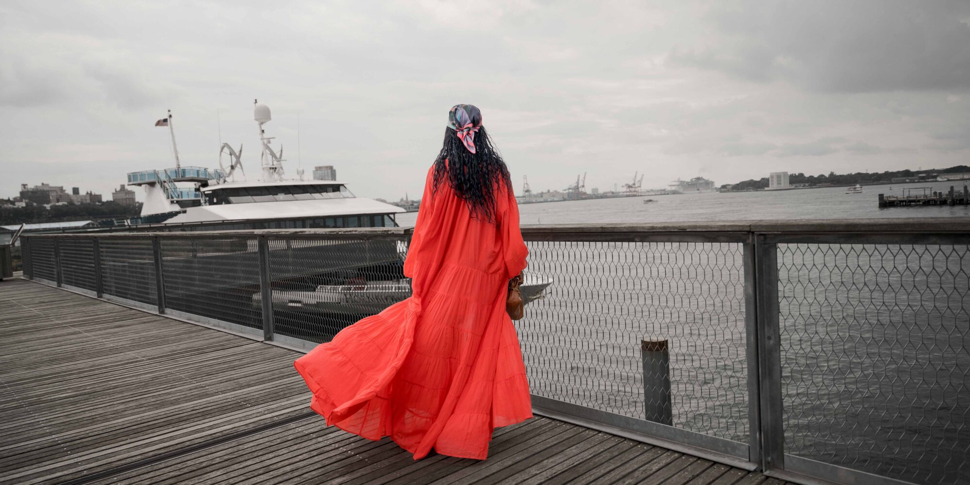 Woman in a red maxi dress poses on a pier at the South Street Seaport in New York City with a view of the skyline and water.