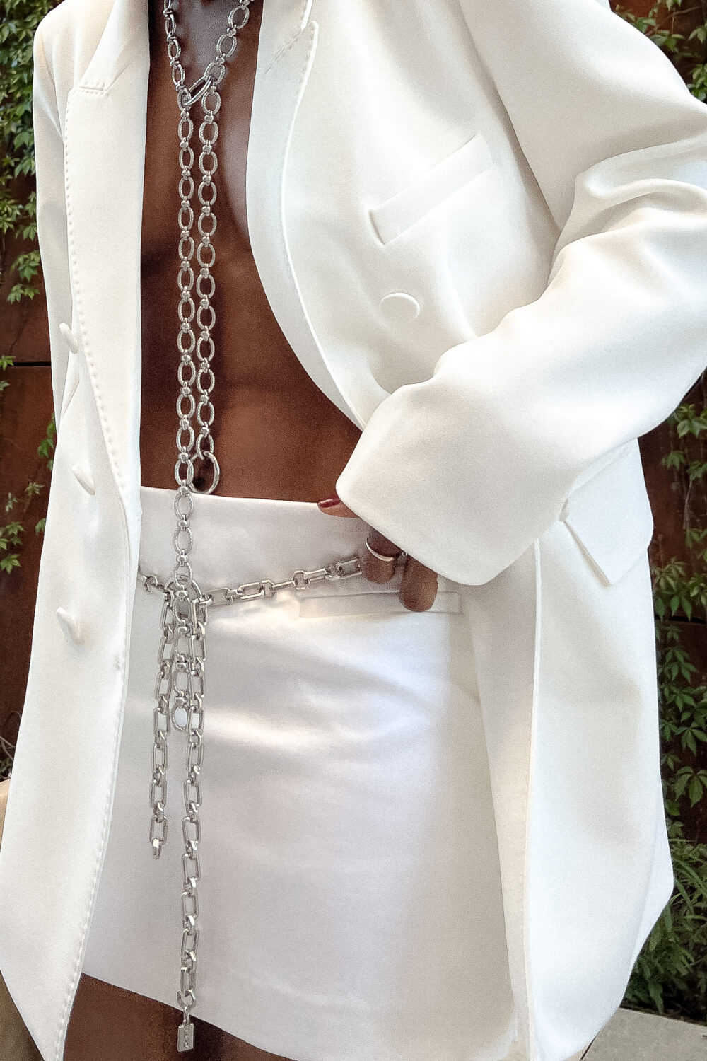 Woman wearing a white mini suit with a floral headband, silver chain belt, red shoes, and a shoulder bag. Modern, stylish outfit.