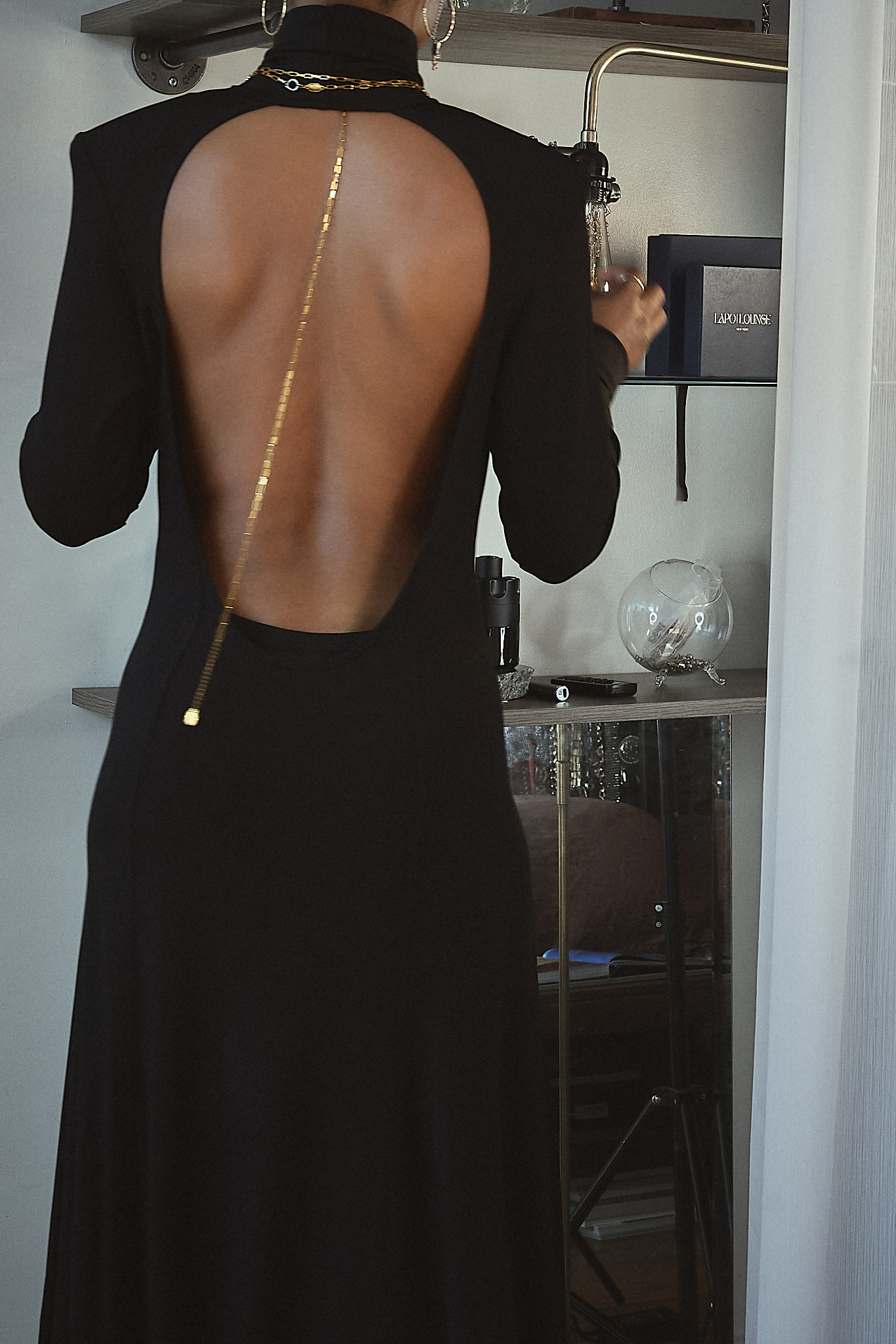 A woman with dark braided hair styled in a low bun poses in a black backless maxi dress with a high neckline and long sleeves. She wears bold gold hoop earrings and an elegant gold chain draped along her exposed back, adding a touch of sophistication. Her poised expression and graceful posture exude confidence and timeless elegance against a neutral background.