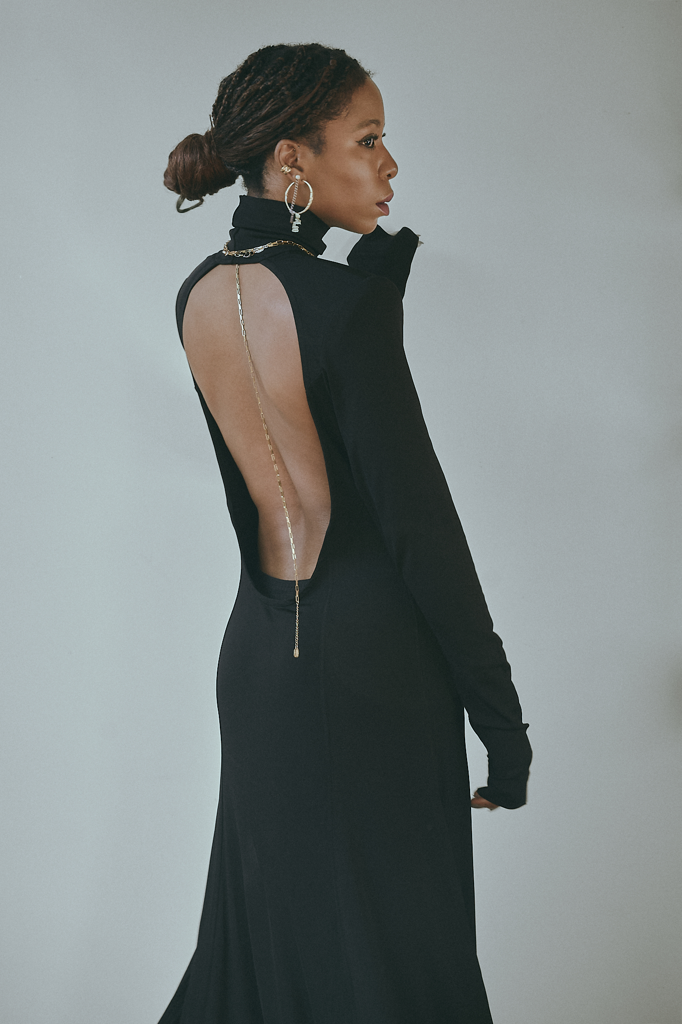 A woman with dark braided hair styled in a low bun poses in a black backless maxi dress with a high neckline and long sleeves. She wears bold gold hoop earrings and an elegant gold chain draped along her exposed back, adding a touch of sophistication. Her poised expression and graceful posture exude confidence and timeless elegance against a neutral background.