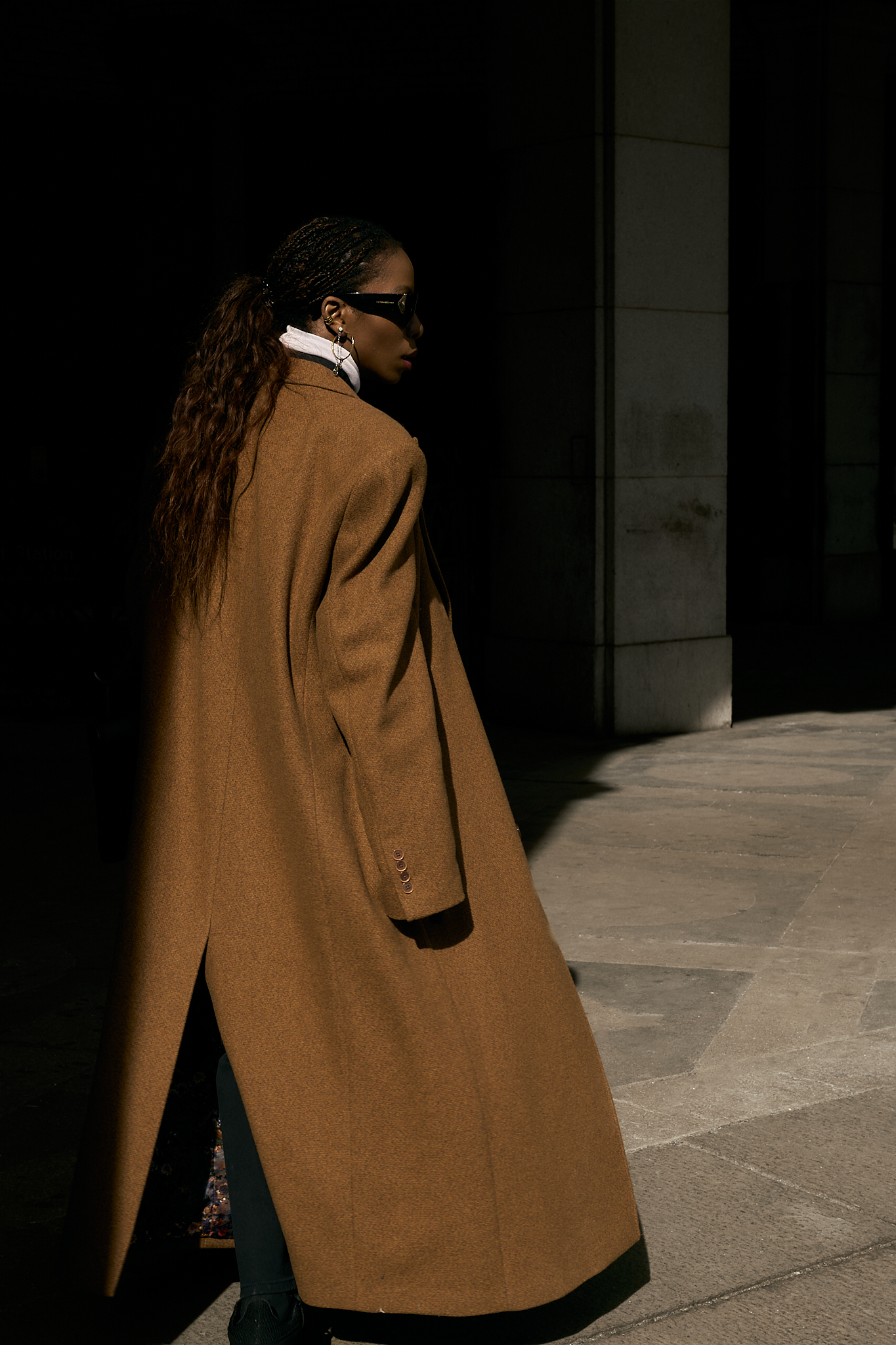 She wears a chic minimalist outfit. A black button-down shirt layered over a crisp white turtleneck, creating a sleek and structured base. A black denim maxi skirt with a front slit adds dimension and movement, while a silver chain belt from Lapo Lounge injects a bold, modern edge. The ensemble is topped with a classic camel coat, adding warmth and sophistication to the monochromatic foundation. Accessories complete the look—oversized black sunglasses for a cool, mysterious vibe, gold hoop earrings for a hint of elegance, and a black leather clutch for a polished finish. She grounds the outfit with black and white sneakers, effortlessly blending high fashion with street-style ease.