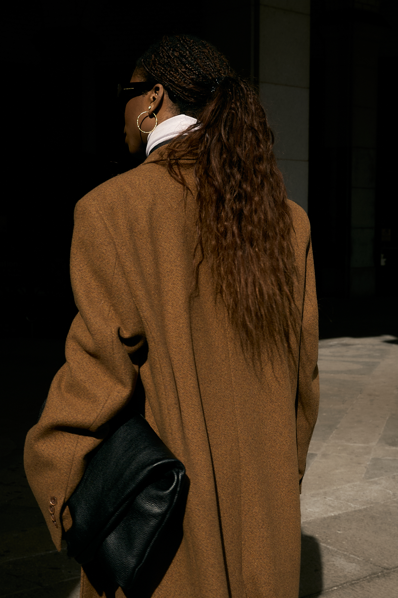 She wears a chic minimalist outfit. A black button-down shirt layered over a crisp white turtleneck, creating a sleek and structured base. A black denim maxi skirt with a front slit adds dimension and movement, while a silver chain belt from Lapo Lounge injects a bold, modern edge. The ensemble is topped with a classic camel coat, adding warmth and sophistication to the monochromatic foundation. Accessories complete the look—oversized black sunglasses for a cool, mysterious vibe, gold hoop earrings for a hint of elegance, and a black leather clutch for a polished finish. She grounds the outfit with black and white sneakers, effortlessly blending high fashion with street-style ease.
