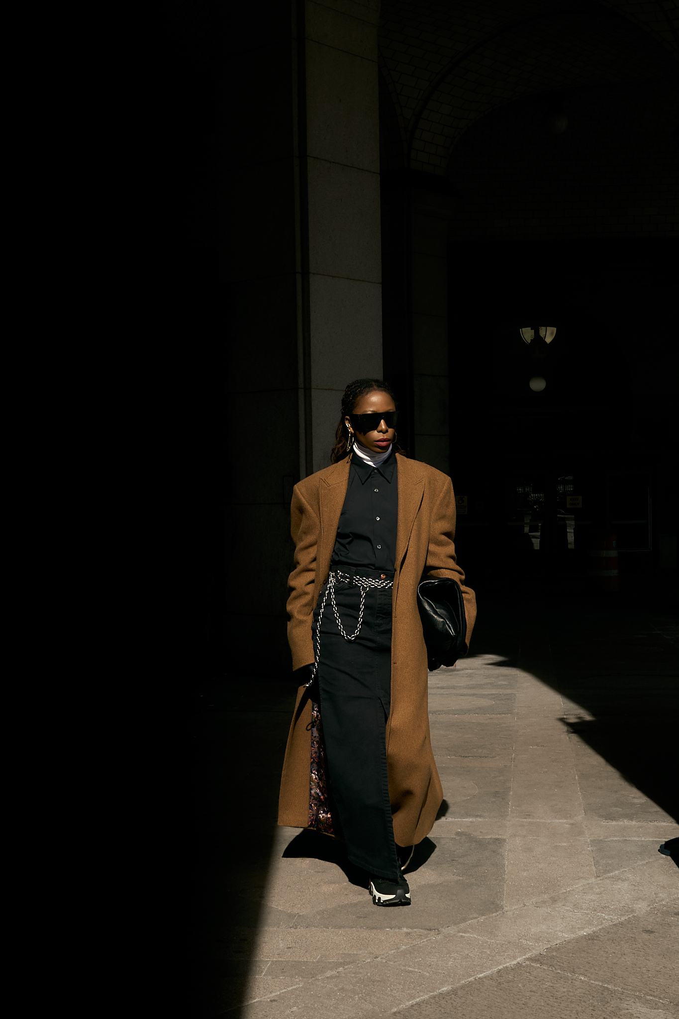 She wears a chic minimalist outfit. A black button-down shirt layered over a crisp white turtleneck, creating a sleek and structured base. A black denim maxi skirt with a front slit adds dimension and movement, while a silver chain belt from Lapo Lounge injects a bold, modern edge. The ensemble is topped with a classic camel coat, adding warmth and sophistication to the monochromatic foundation. Accessories complete the look—oversized black sunglasses for a cool, mysterious vibe, gold hoop earrings for a hint of elegance, and a black leather clutch for a polished finish. She grounds the outfit with black and white sneakers, effortlessly blending high fashion with street-style ease.
