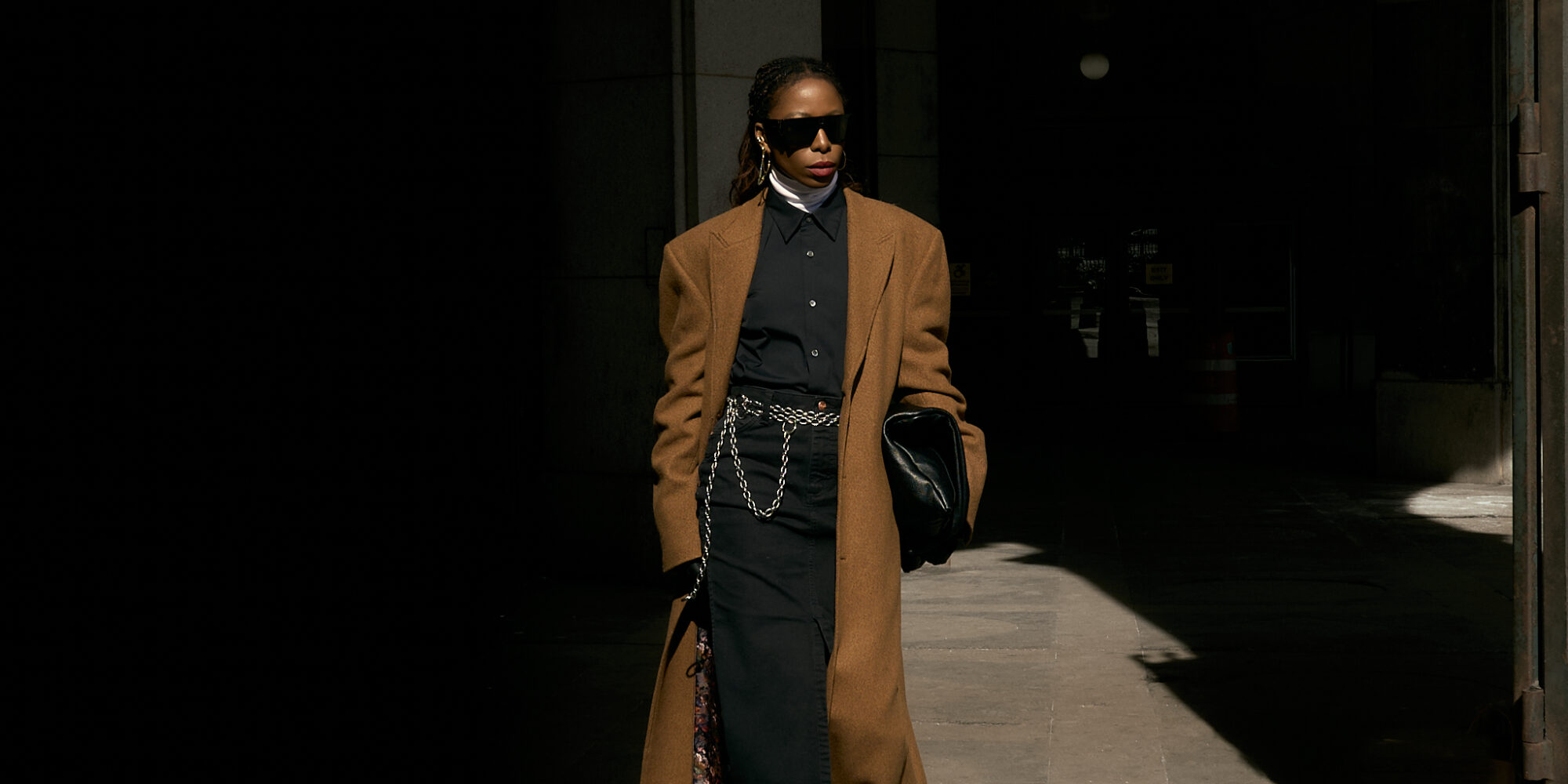 She wears a black button-down shirt layered over a crisp white turtleneck, creating a sleek and structured base. A black denim maxi skirt with a front slit adds dimension and movement, while a silver chain belt from Lapo Lounge injects a bold, modern edge. The ensemble is topped with a classic camel coat, adding warmth and sophistication to the monochromatic foundation. Accessories complete the look—oversized black sunglasses for a cool, mysterious vibe, gold hoop earrings for a hint of elegance, and a black leather clutch for a polished finish. She grounds the outfit with black and white sneakers, effortlessly blending high fashion with street-style ease.