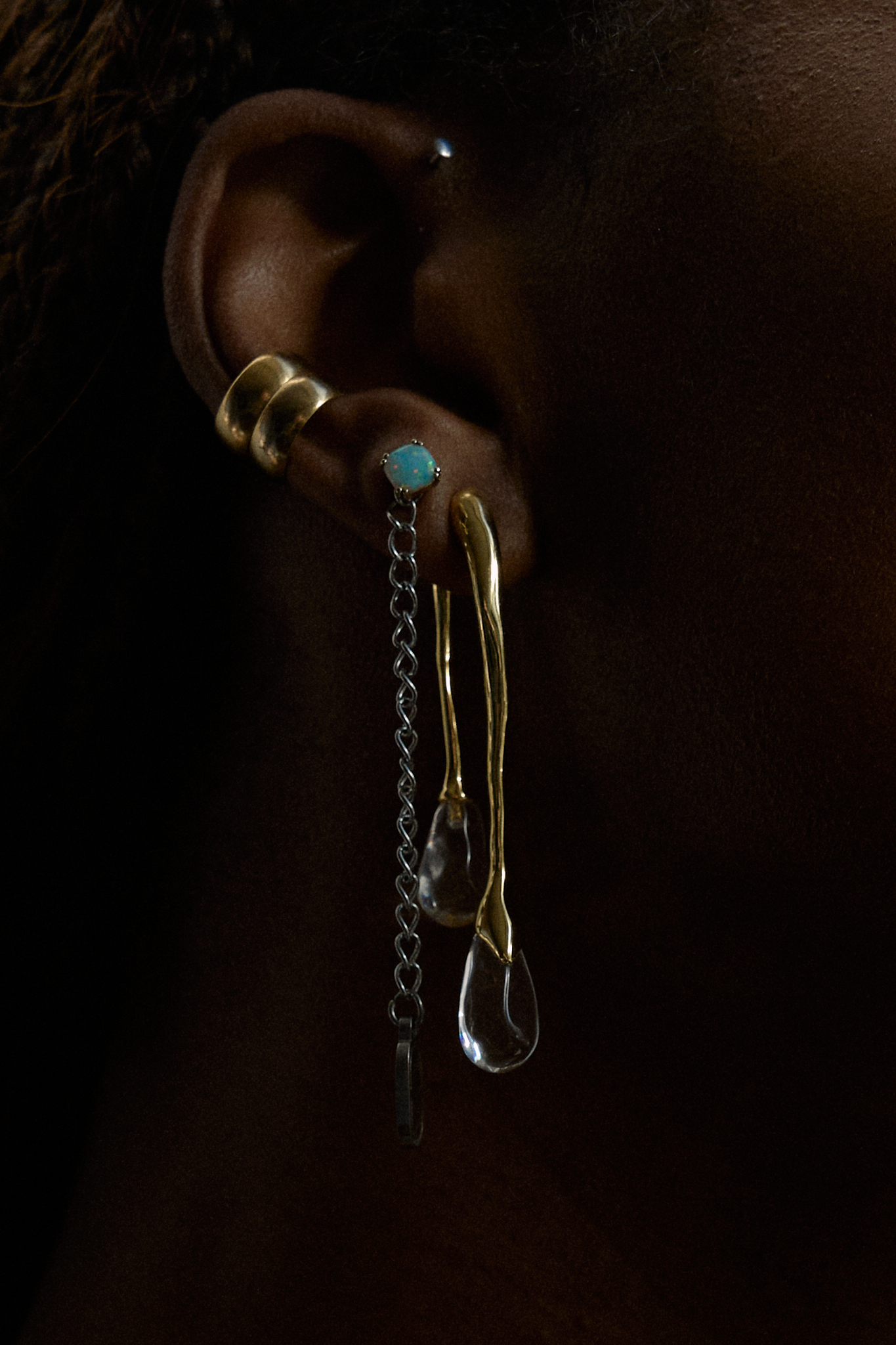 Close-up of model wearing a minimalist opal stud earring, Alexis Bittar clear lucite drop earring, and two layered gold ear cuffs — a modern mix of statement and delicate jewelry for a chic, everyday look.