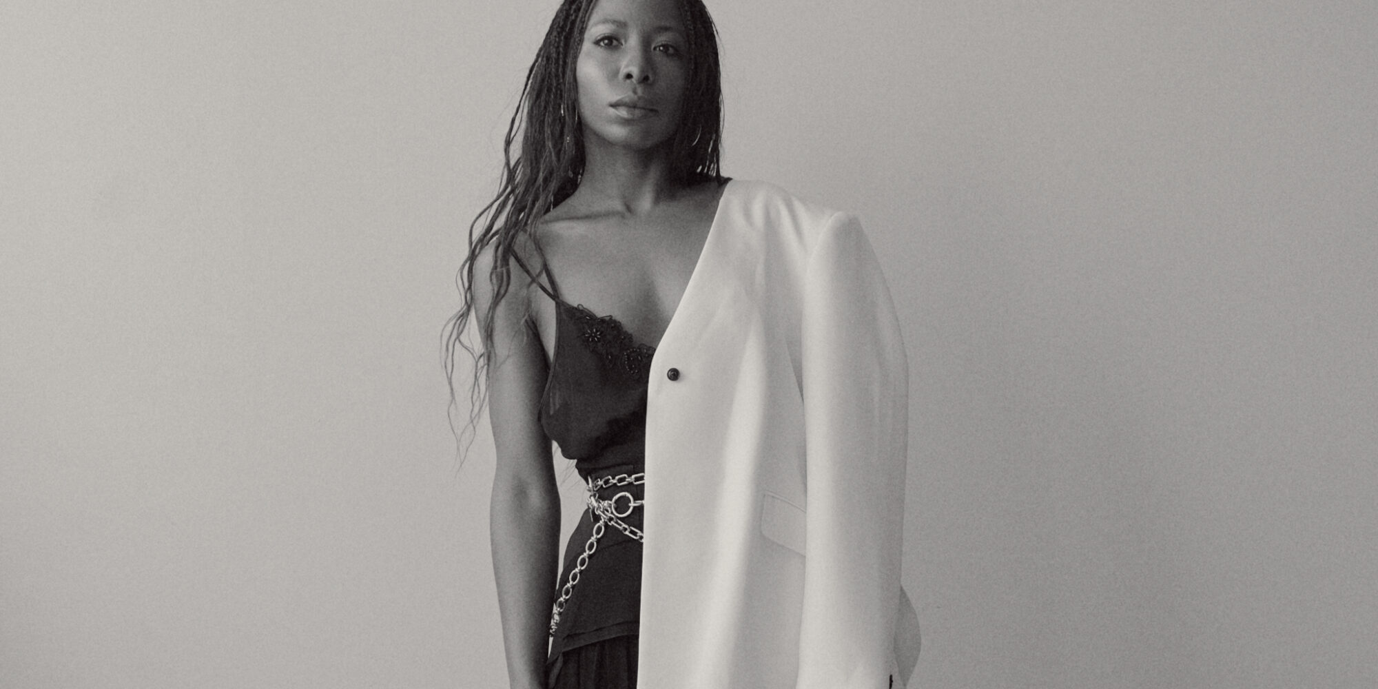 Woman wearing an oversized white blazer paired with a black flowy maxi skirt, styled with layered silver chain belts and silver necklaces, standing confidently against a modern white backdrop.