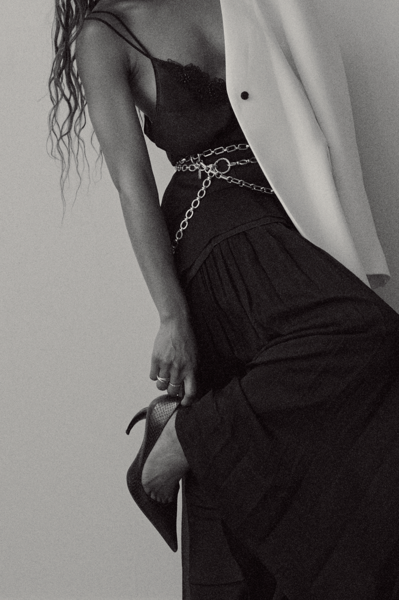 Woman wearing an oversized white blazer paired with a black flowy maxi skirt, styled with layered silver chain belts and silver necklaces, standing confidently against a modern white backdrop.