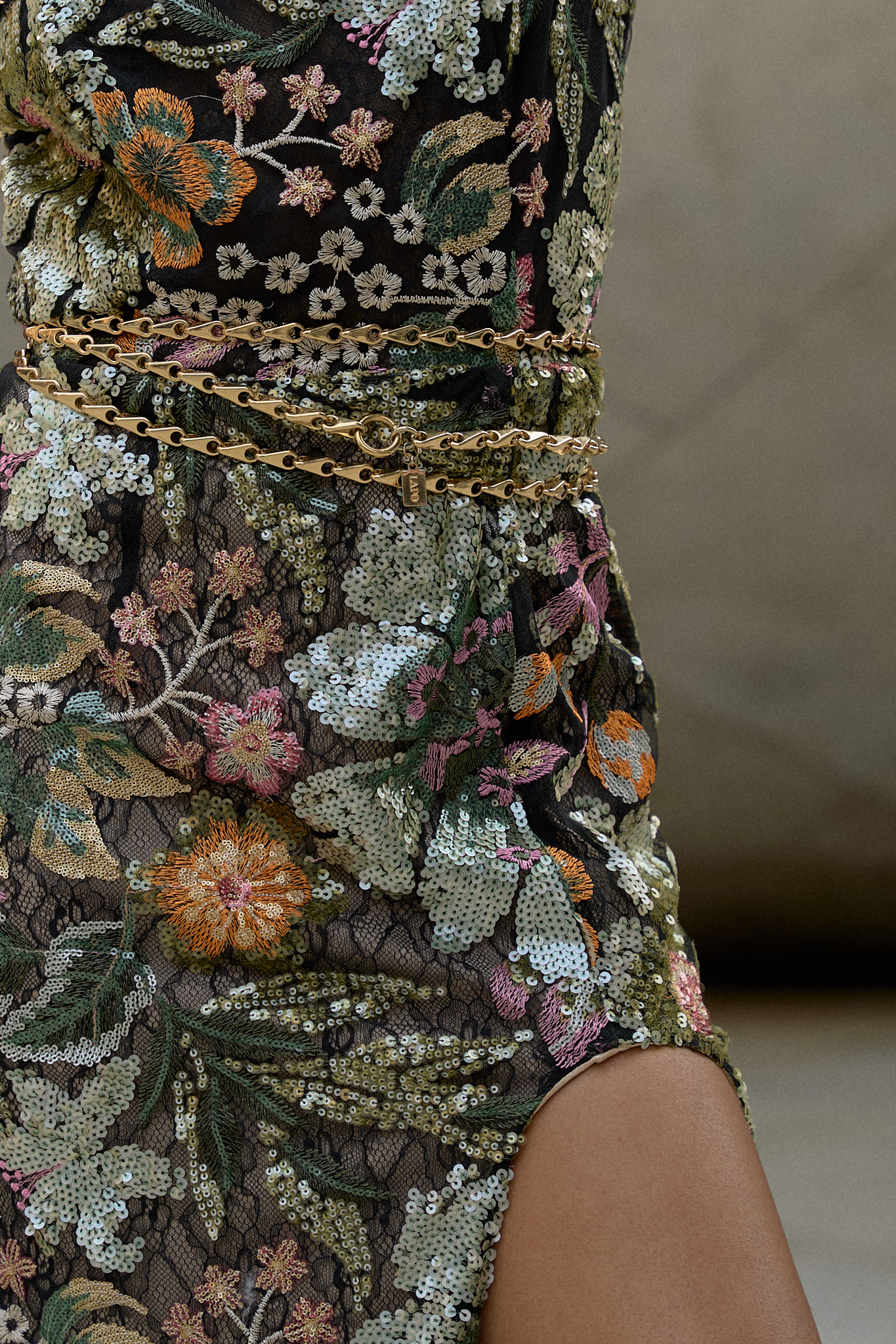 Close-up of a floral sequined wedding guest dress featuring intricate embroidery and a gold chain belt, held by a hand adorned with rings and bold red nail polish.