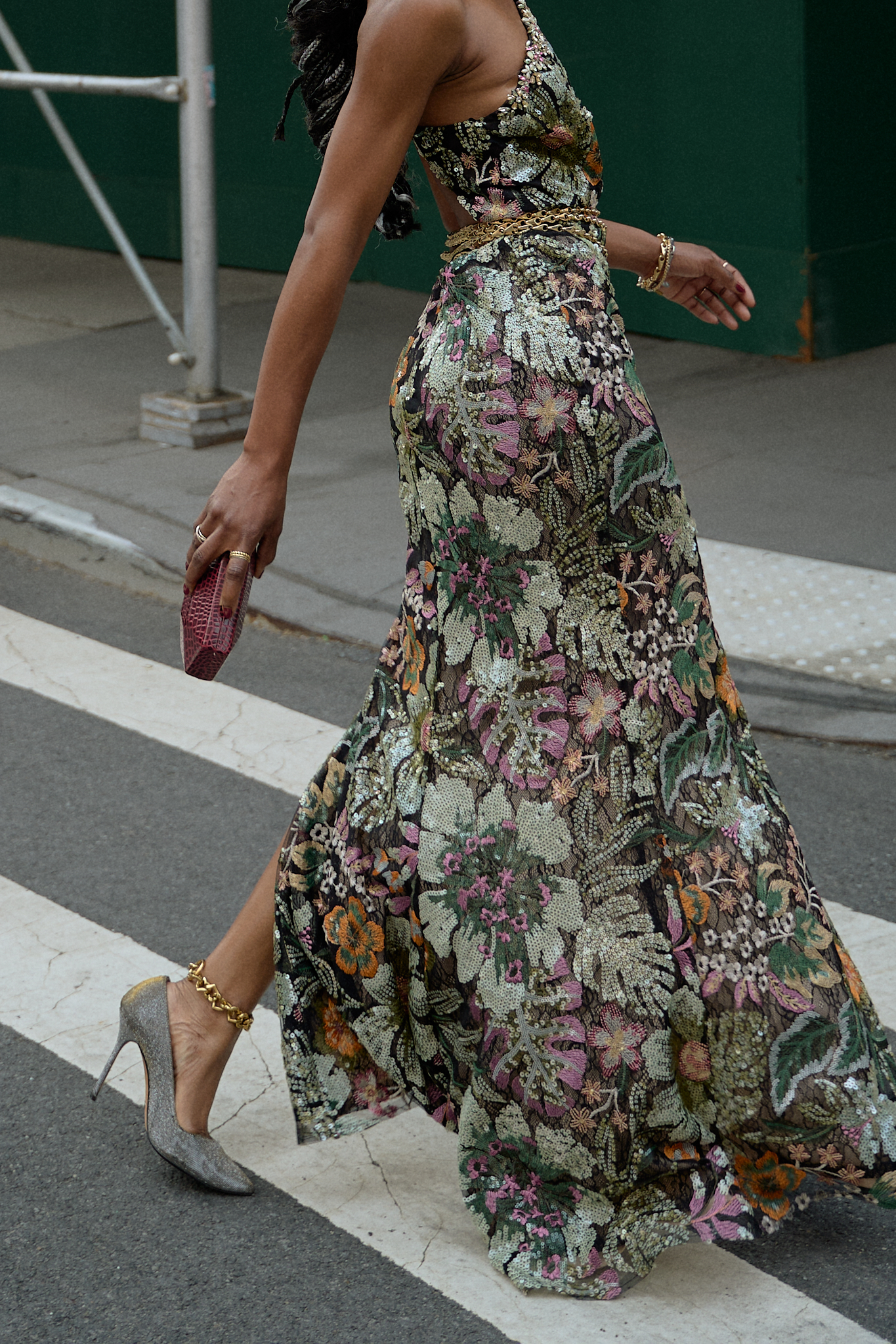 A stylish woman walking on a street in New York City in a floral patterned maxi dress with a high slit and a back cutout, holding a small red clutch and wearing gold chain belt and silver heels.