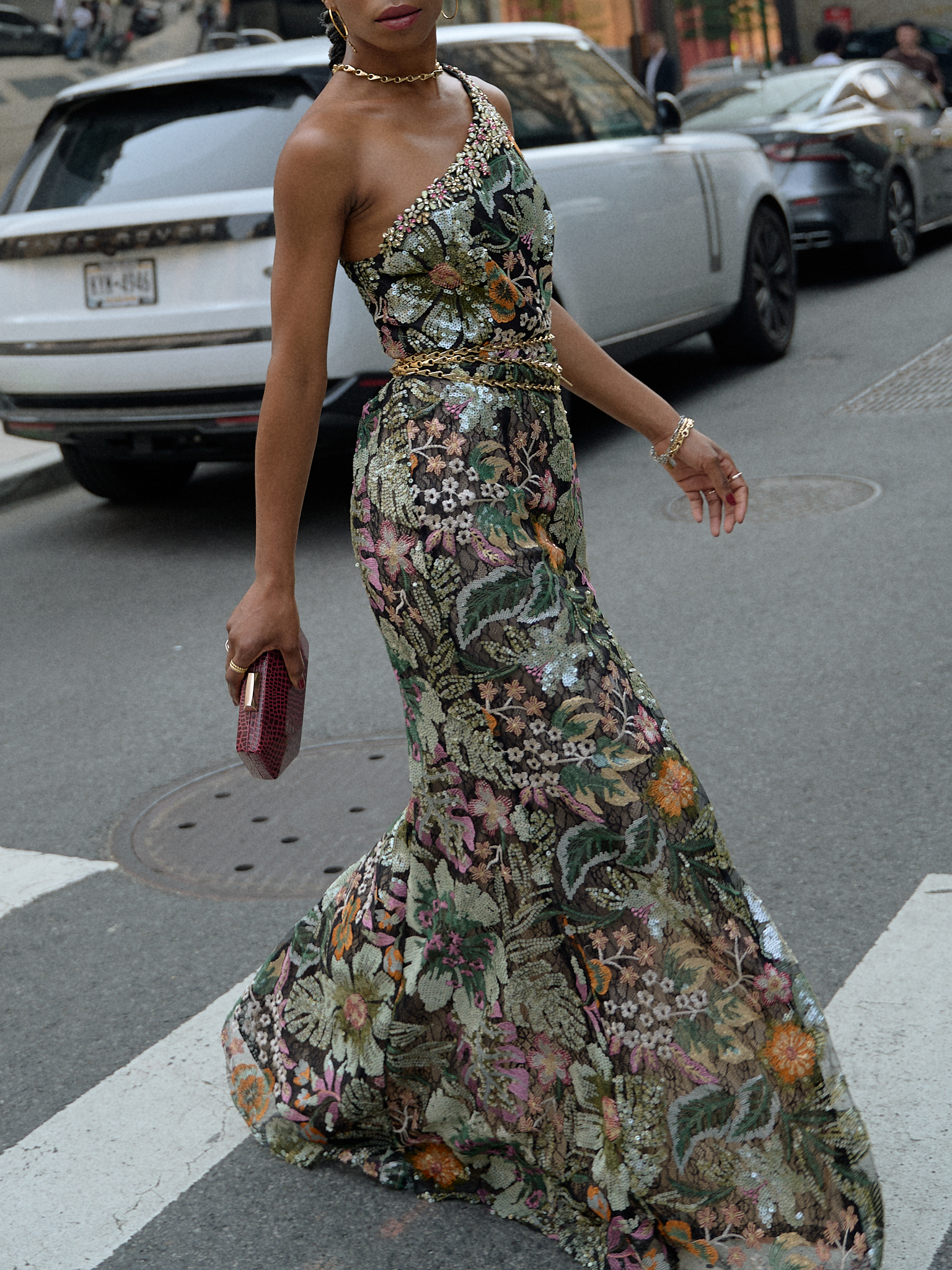 A stylish woman walking in a floral, sequined gown with one shoulder exposed, paired with gold chain belt and gold necklace, in New York City.