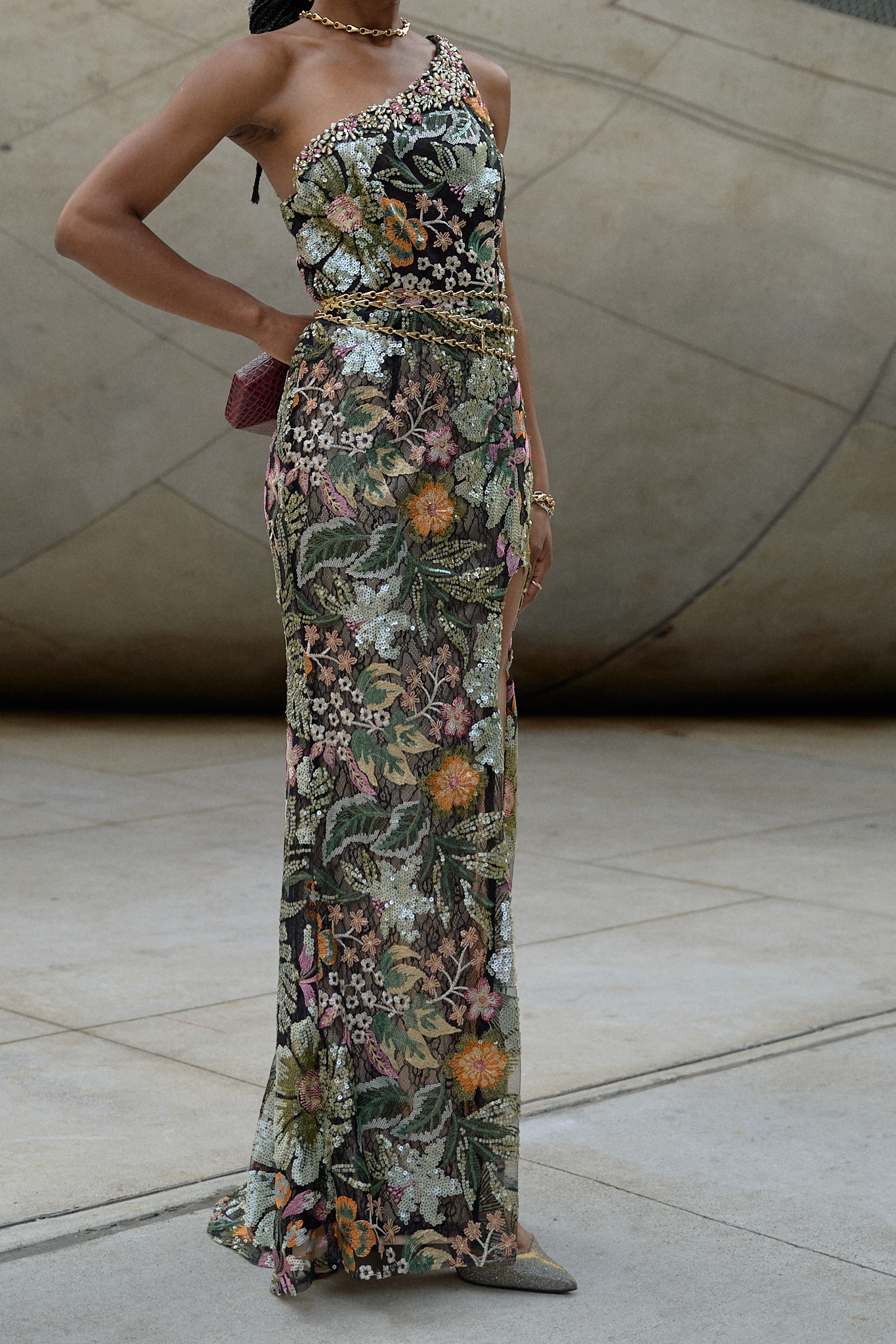 A stylish woman wearing a floral sequin evening gown with one shoulder and a thigh-high slit, styled with a gold chain belt and holding a small bag, standing outdoors against a textured concrete background.