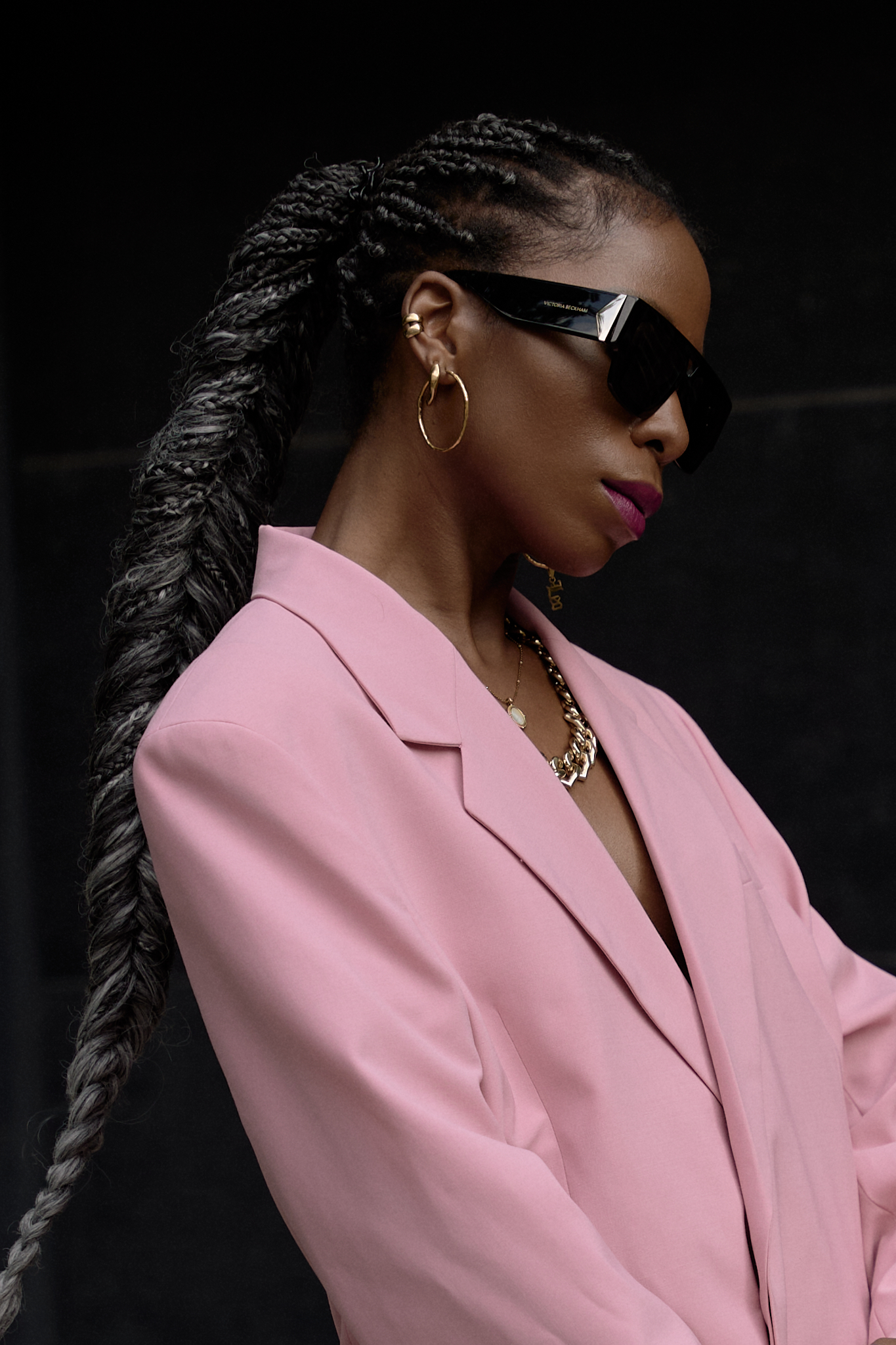 Woman wearing a structured oversized pink blazer and matching asymmetrical mini skirt, styled with chunky black sneakers, gold chain necklace, hoop earrings, black sunglasses, and a pink handbag with a gold chain strap, standing confidently on a city street.