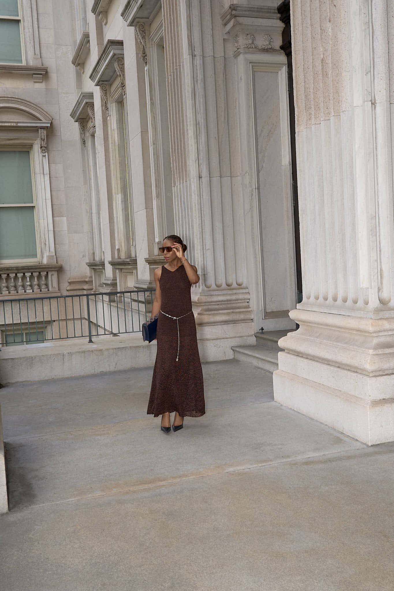 A woman wearing a sleeveless,brown maxi knit dress. The dress is cinched at the waist with a simple lapo lounge silver chain belt. She is also carrying a navy blue clutch and wearing black stiletto heels, sunglasses, and delicate silver jewelry. The background is a large, classical building with stone columns in FiDi NYC.