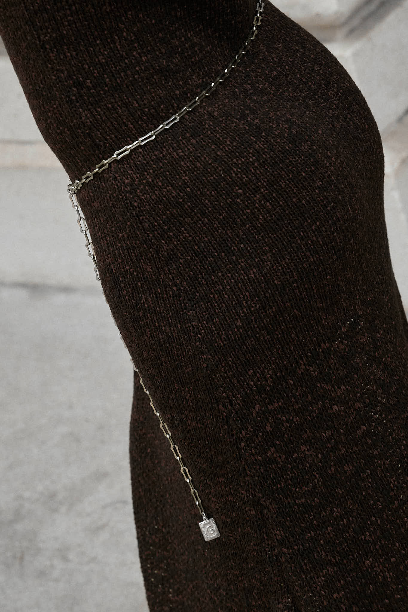 A woman wearing a sleeveless,brown maxi knit dress. The dress is cinched at the waist with a simple lapo lounge silver chain belt. She is also carrying a navy blue clutch and wearing black stiletto heels, sunglasses, and delicate silver jewelry. The background is a large, classical building with stone columns in FiDi NYC.