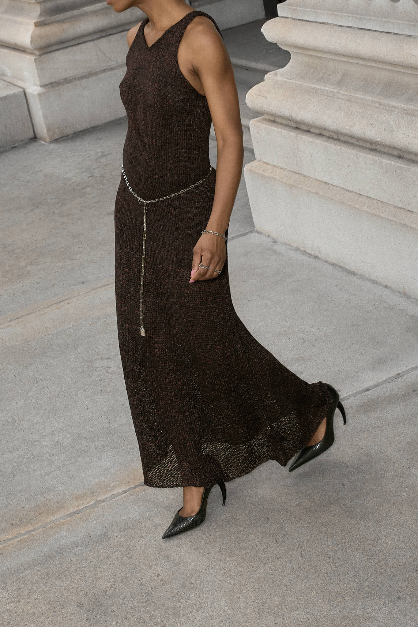 A woman wearing a sleeveless,brown maxi knit dress. The dress is cinched at the waist with a simple lapo lounge silver chain belt. She is also carrying a navy blue clutch and wearing black stiletto heels, sunglasses, and delicate silver jewelry. The background is a large, classical building with stone columns in FiDi NYC.