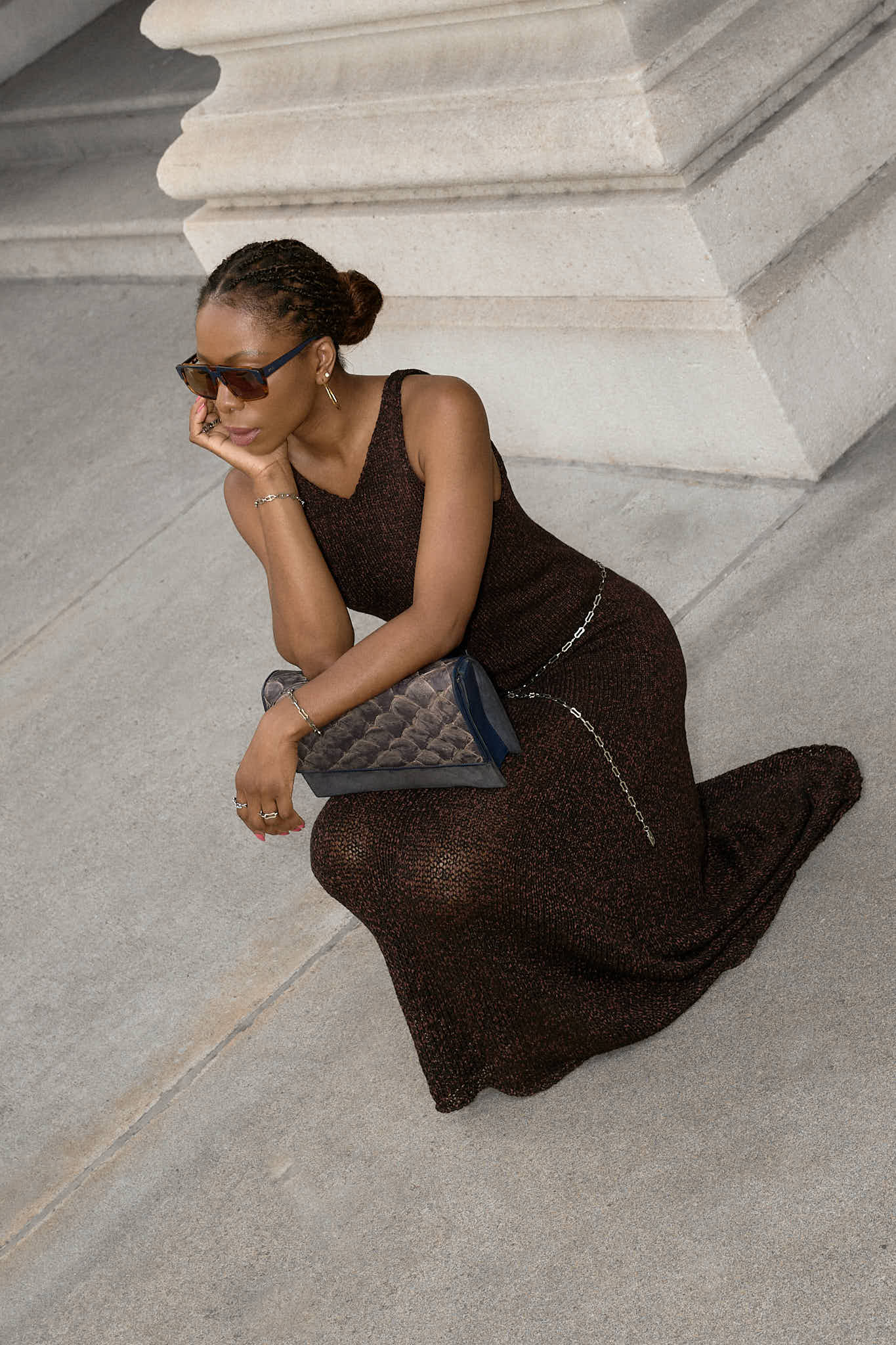 A woman wearing a sleeveless,brown maxi knit dress. The dress is cinched at the waist with a simple lapo lounge silver chain belt. She is also carrying a navy blue clutch and wearing black stiletto heels, sunglasses, and delicate silver jewelry. The background is a large, classical building with stone columns in FiDi NYC.