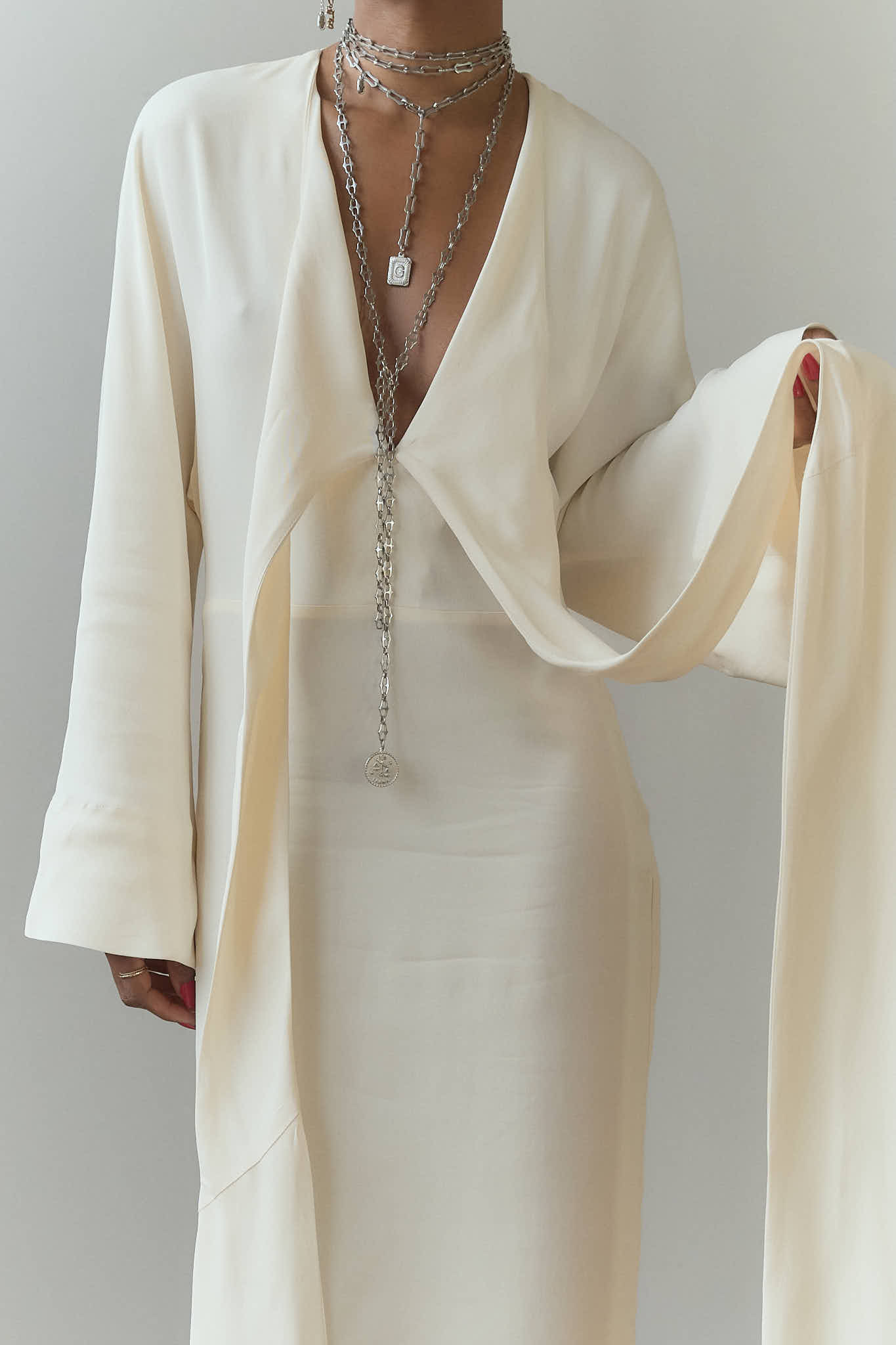 A close-up of a person wearing a white long-sleeve maxi dress, styled with layered Lapo Lounge silver chunky link necklaces including a long lariat chain with a coin pendant.