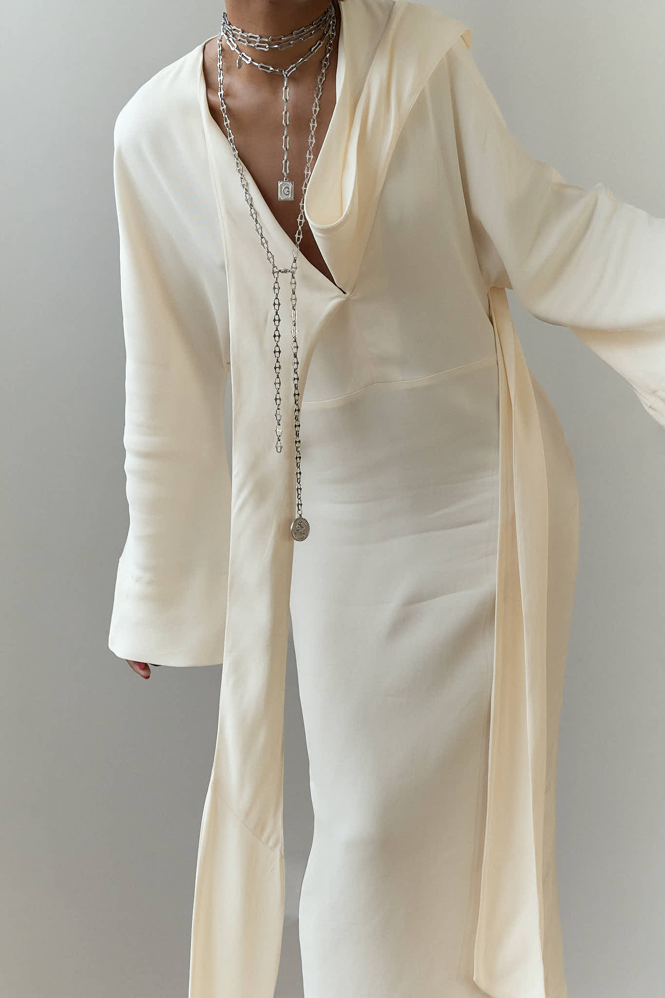 Mid-shot of layered silver lariat and chunky link necklaces on a cream maxi dress.