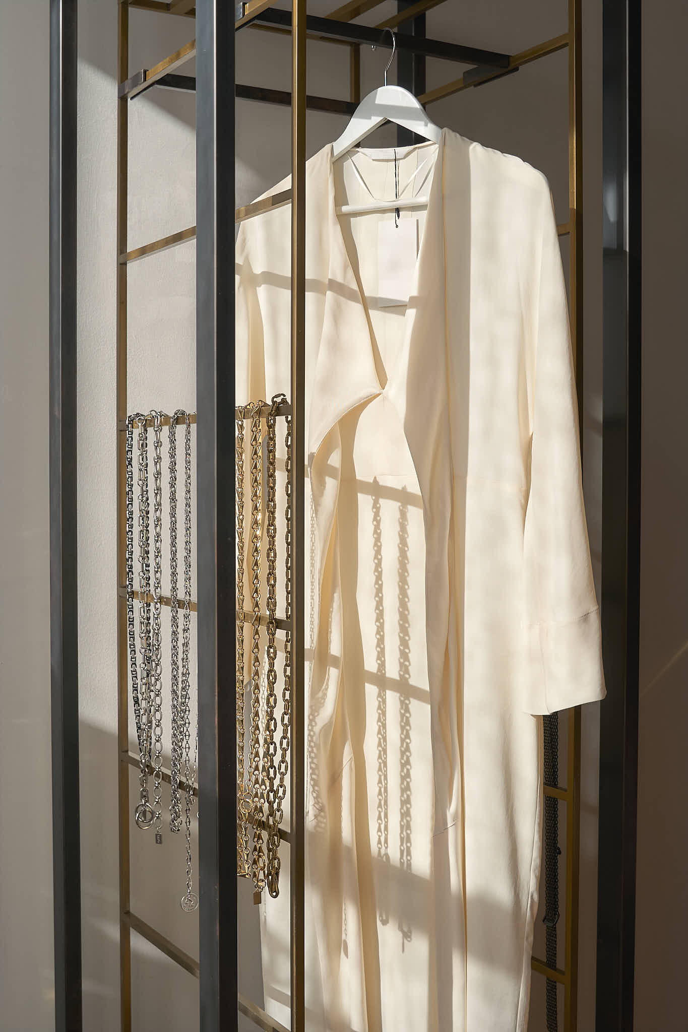 A white long-sleeve dress hanging on a white hanger inside a gold-framed display rack, with silver and gold chunky link chain belts hanging neatly to the left.