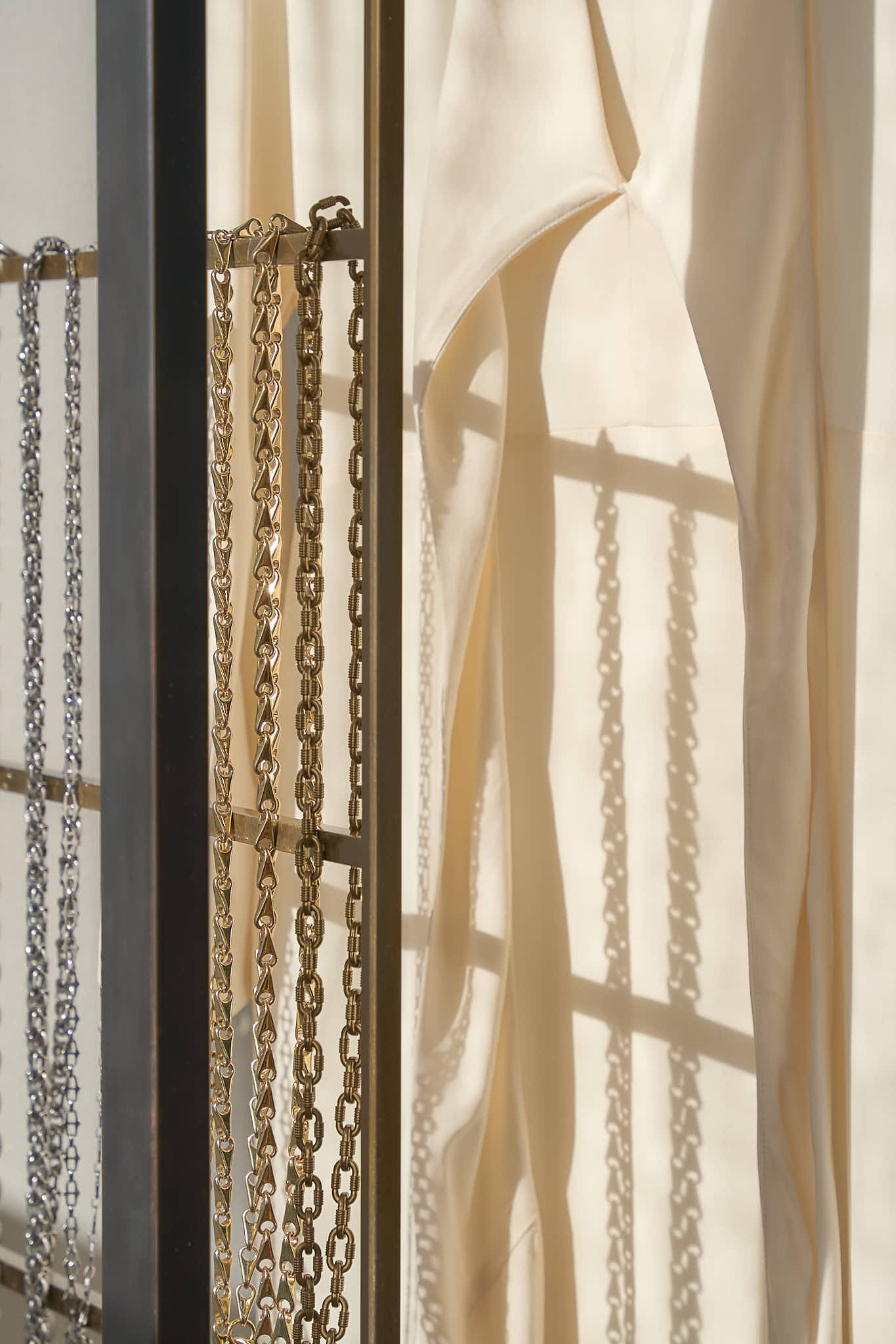 Detail shot of silver and gold link chain belts hanging on a metal display rack, with the soft shadows of a white dress cast against the wall in the background.