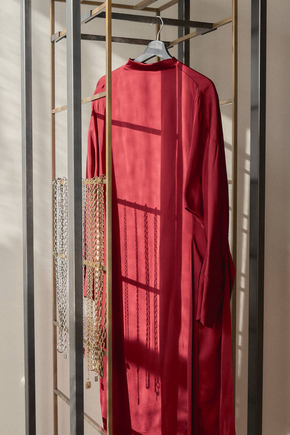 Red satin maxi dress with open back keyhole detail on hanger, displayed with gold chain belt jewelry on modern clothing rack with afternoon light