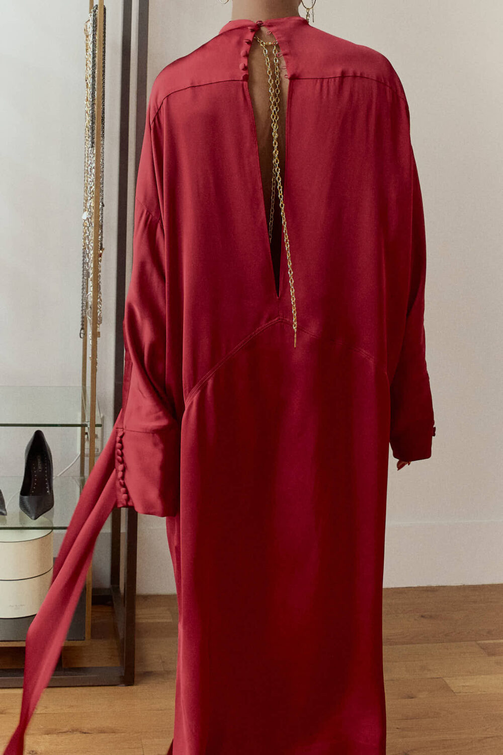 Wine red satin maxi dress back view showing open keyhole back detail styled with gold chain back jewelry, red side ties, and black moto boots