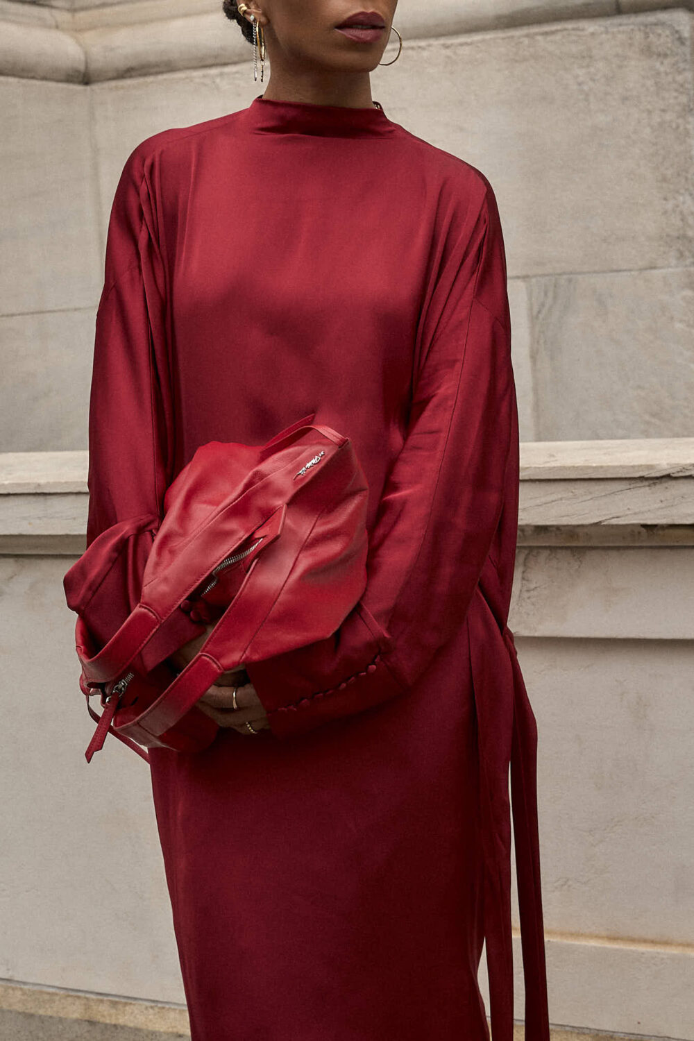 Portrait of a woman in a red silk dress and bold black sunglasses, holding a red leather bag against a new york city background.