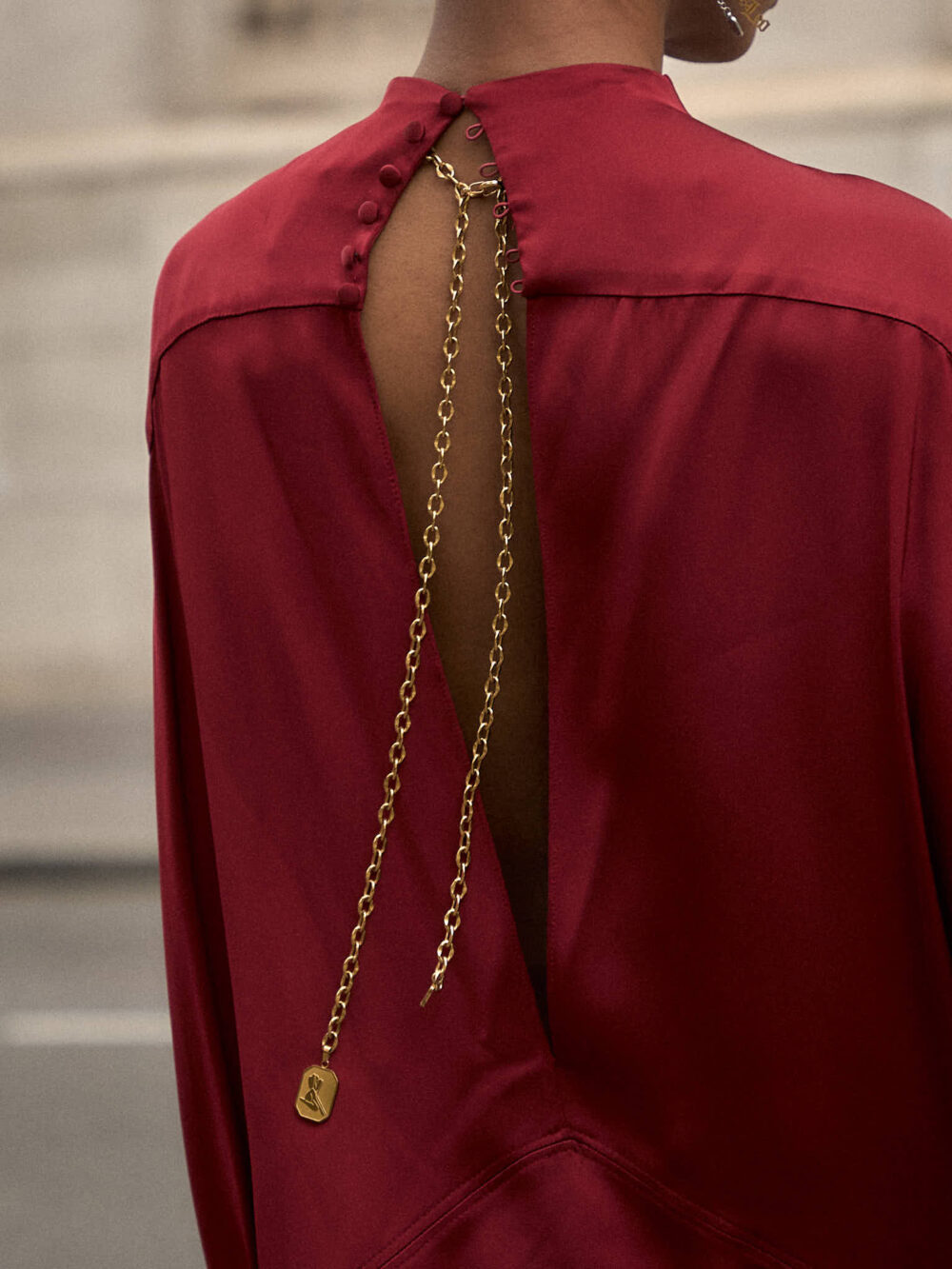 Close-up of the back of a red silk dress, showing a deep slit held together at the top by a thin gold chain that drapes down the bare back.