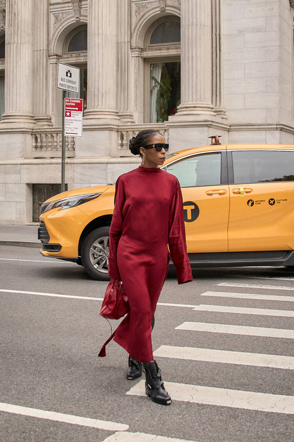 A woman walking in a new york city crosswalk wearing a monochromatic oxblood silk maxi dress with trailing ribbons, styled with black buckle-detail moto boots and a matching red leather bag.