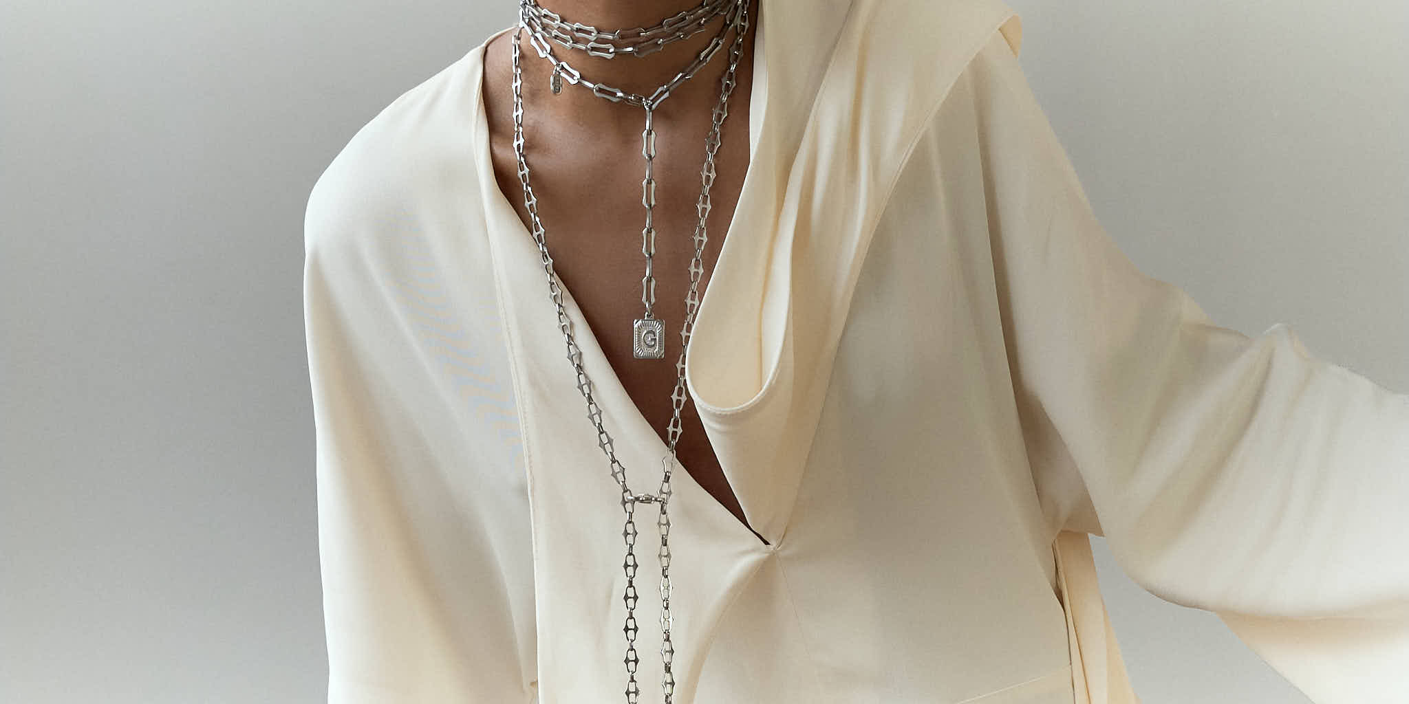 A close-up of a person wearing a white long-sleeve maxi dress, styled with layered Lapo Lounge silver chunky link necklaces including a long lariat chain with a coin pendant.