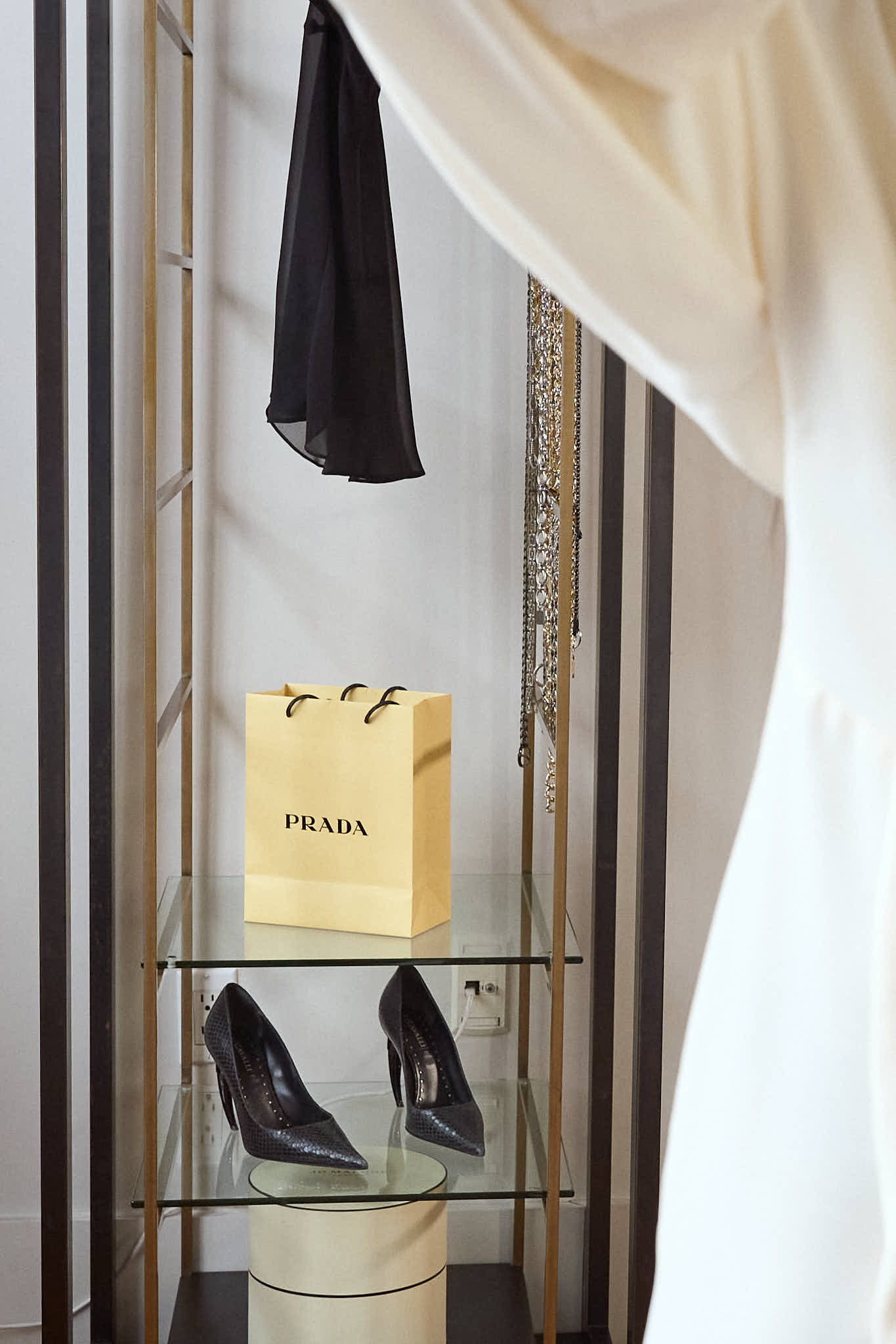Close-up of a minimalist glass shelf featuring a yellow Prada shopping bag and a pair of black pointed-toe stiletto pumps, with jewelry hanging in the background.