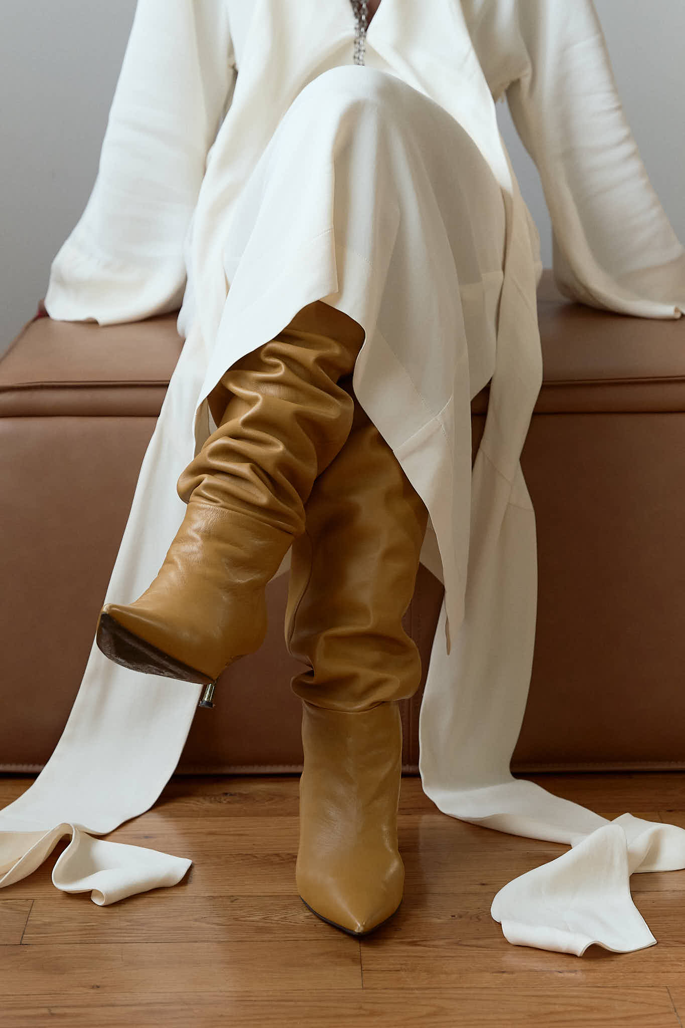 Person seated on an ottoman wearing tan slouchy high-heeled boots and a white dress.