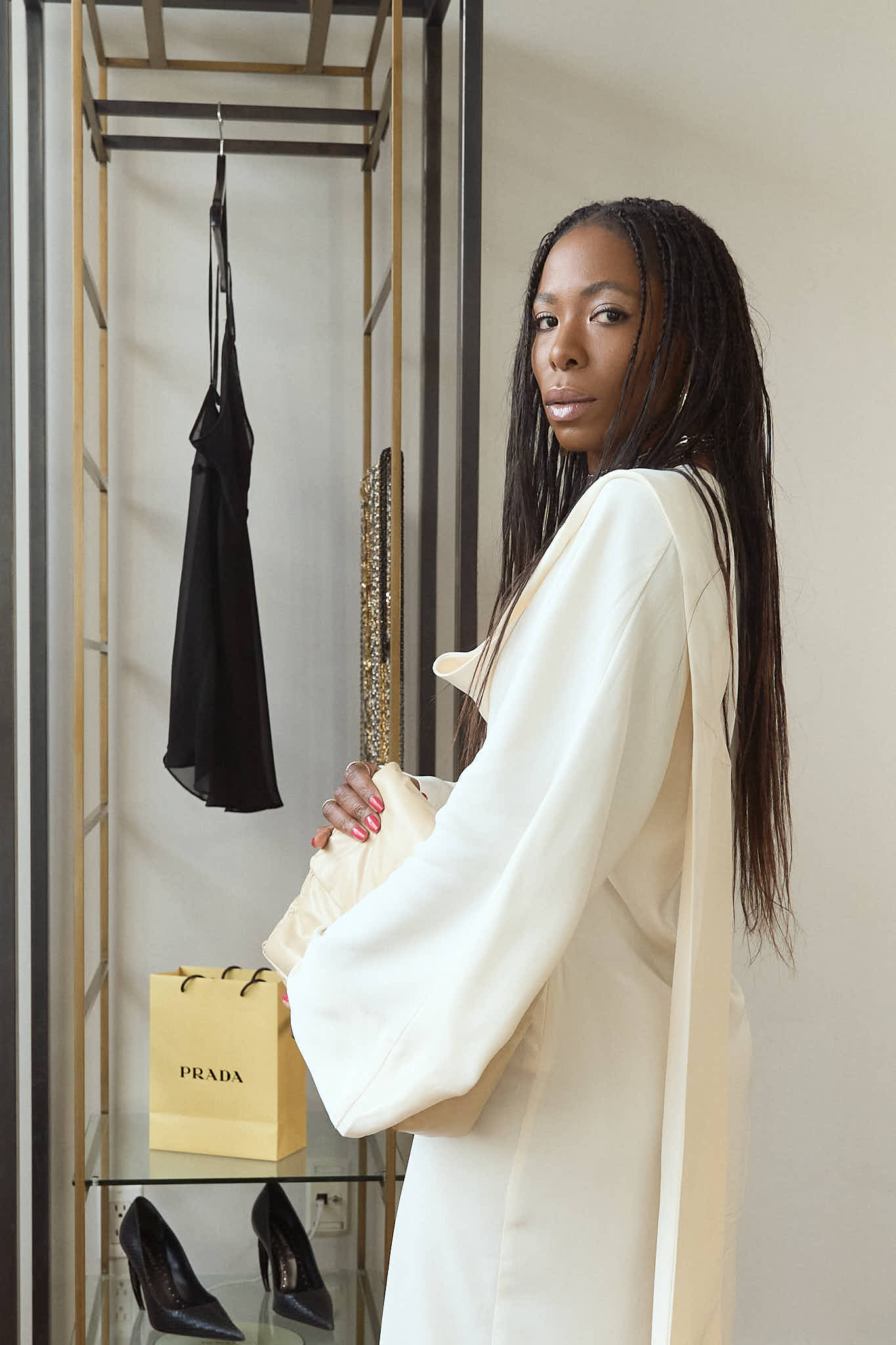 Editorial portrait of a woman with long braids posing in a white maxi dress in front of a modern gold and black garment rack with a Prada shopping bag nearby.