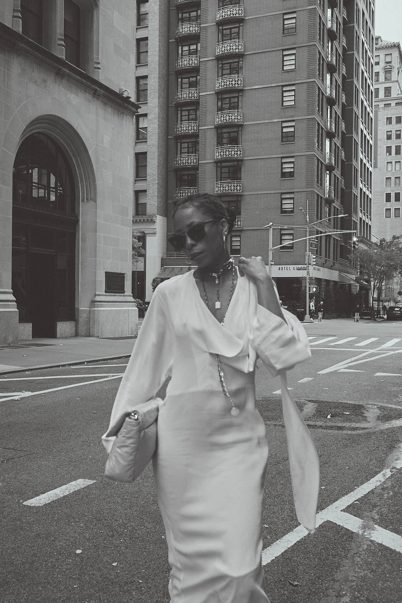 Black and white street style photograph of a woman walking in New York City wearing a long white maxi dress, sunglasses, and multiple silver necklaces, carrying a large clutch bag.