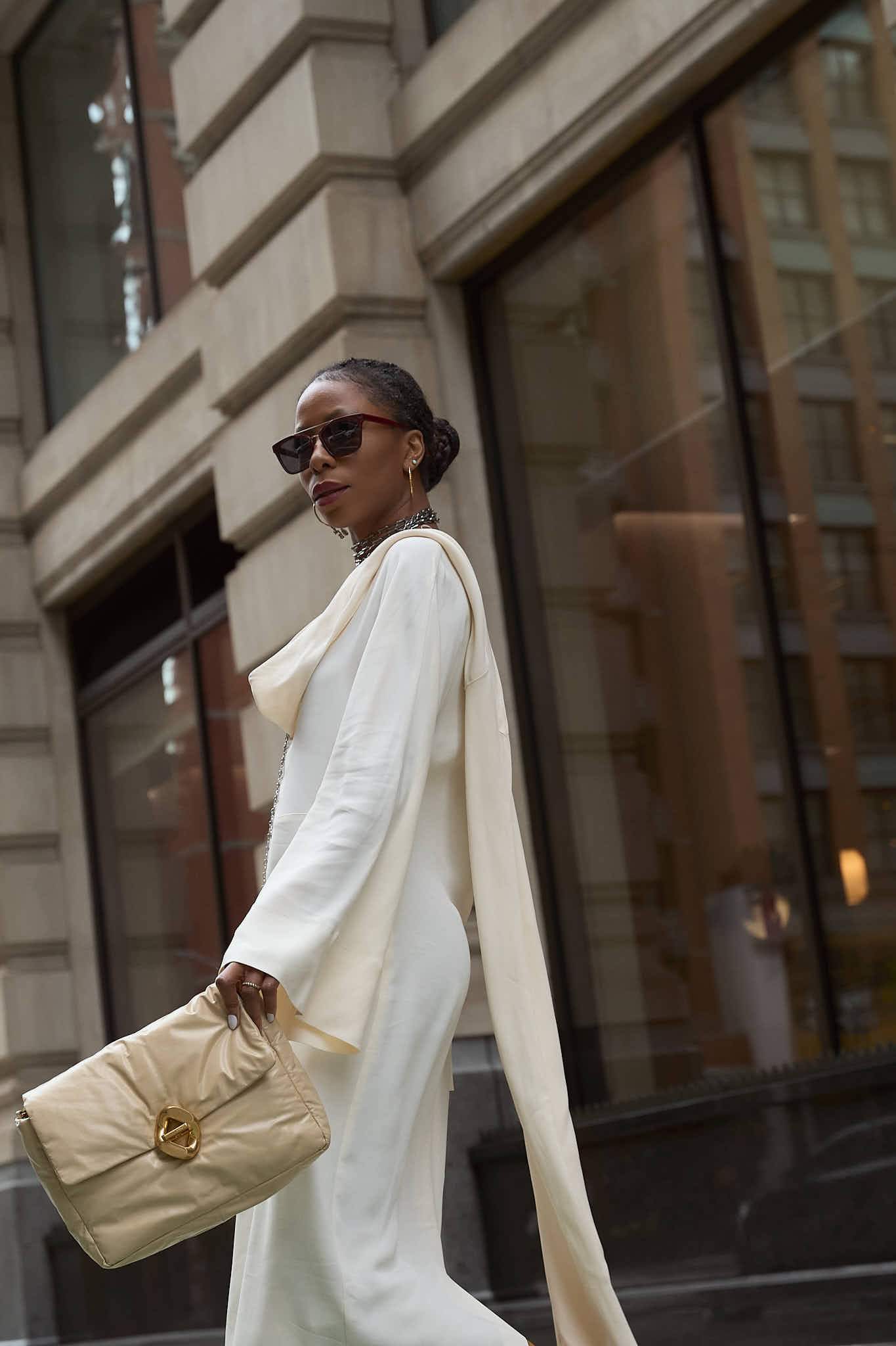 Low angle street photo of a woman in a white dress and sunglasses walking past an urban building.